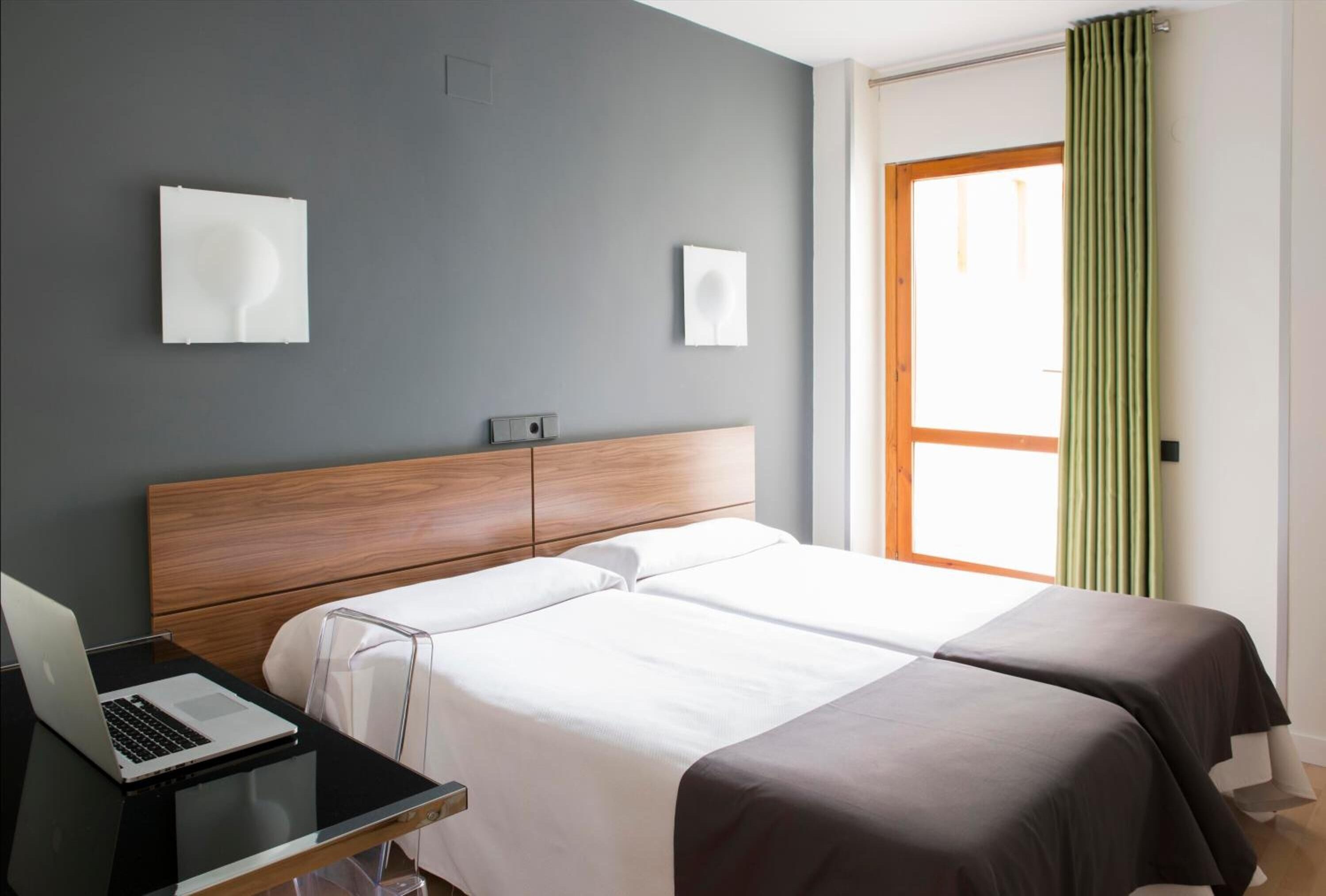 Hotelkamer met twee bedden, een bureau met een laptop, een houten hoofdeinde en een raam met gordijnen.