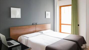 Hotelkamer met twee bedden, een bureau met een laptop, een houten hoofdeinde en een raam met gordijnen.
