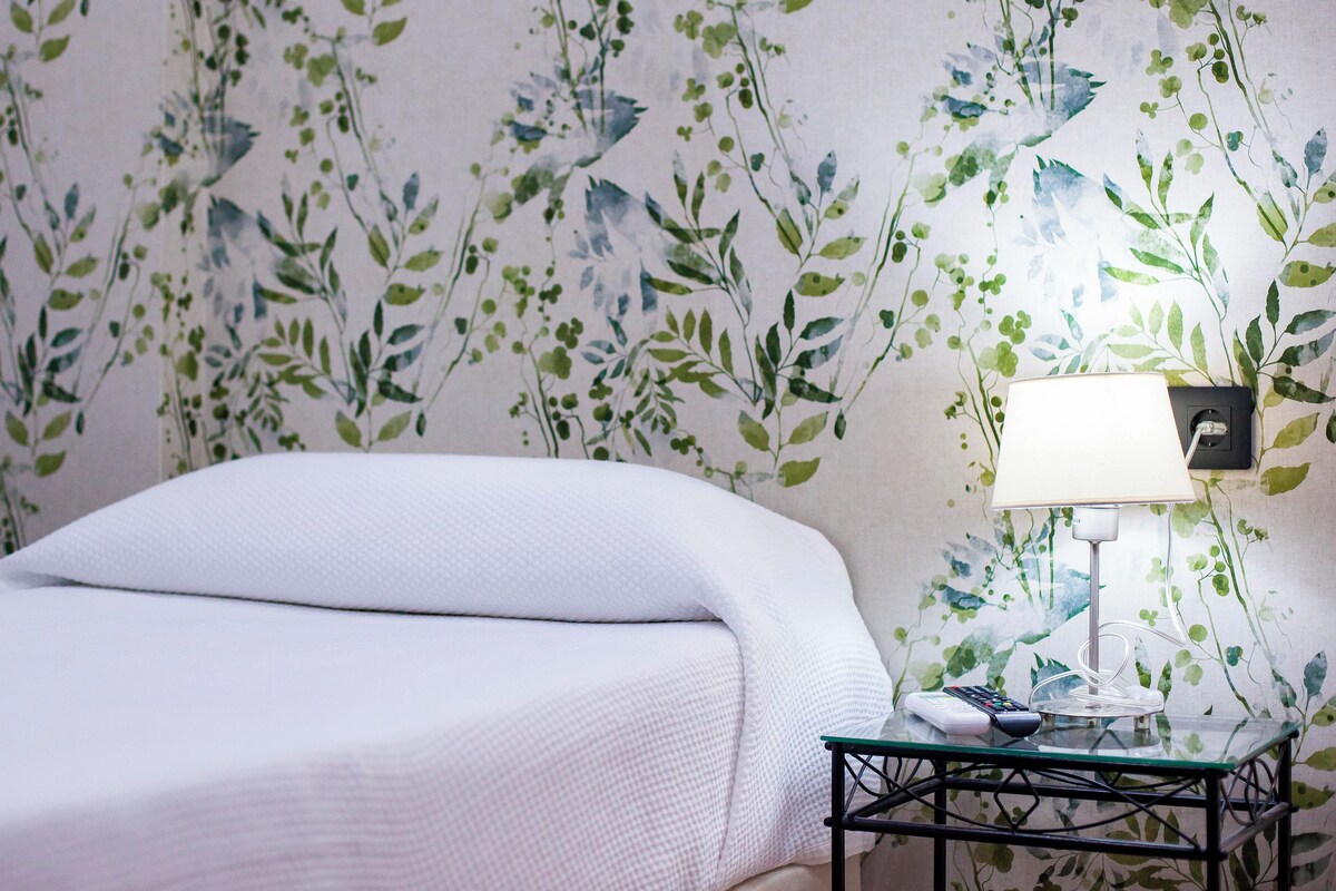 A neatly made bed with a white bedspread, a glass table with a lamp, and a wall with a leafy wallpaper pattern.