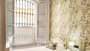 A hotel room with two beds, a window with a metal grill, a nightstand with a lamp, and a wall with a leafy wallpaper pattern. - Hostal San Francisco (Seville)