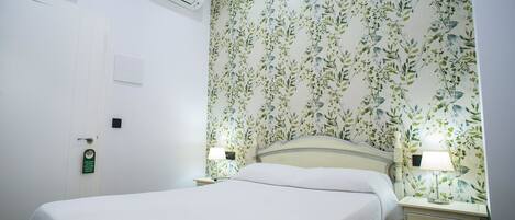 A neatly made bed with a white bedspread, two bedside lamps, a floral wallpaper, a white door with a lock, and an air conditioning unit.