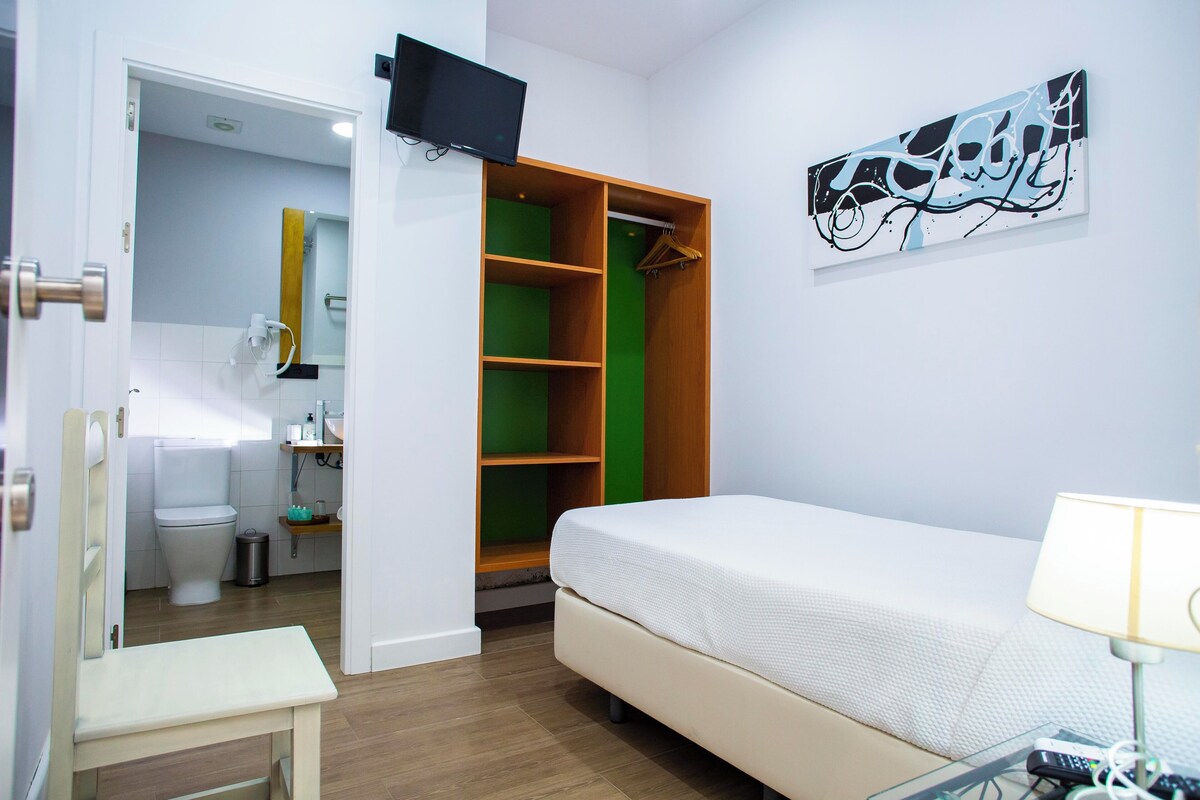 A bedroom with a bed, a bedside table, a TV mounted on the wall, a wardrobe, and a bathroom visible through an open door.