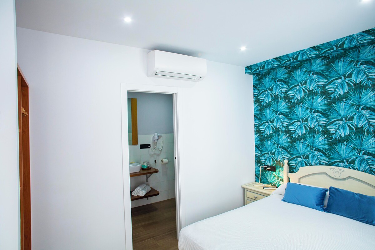 A bedroom with a bed, a nightstand, a wall-mounted air conditioner, a bathroom with a towel rack, and a shower area.