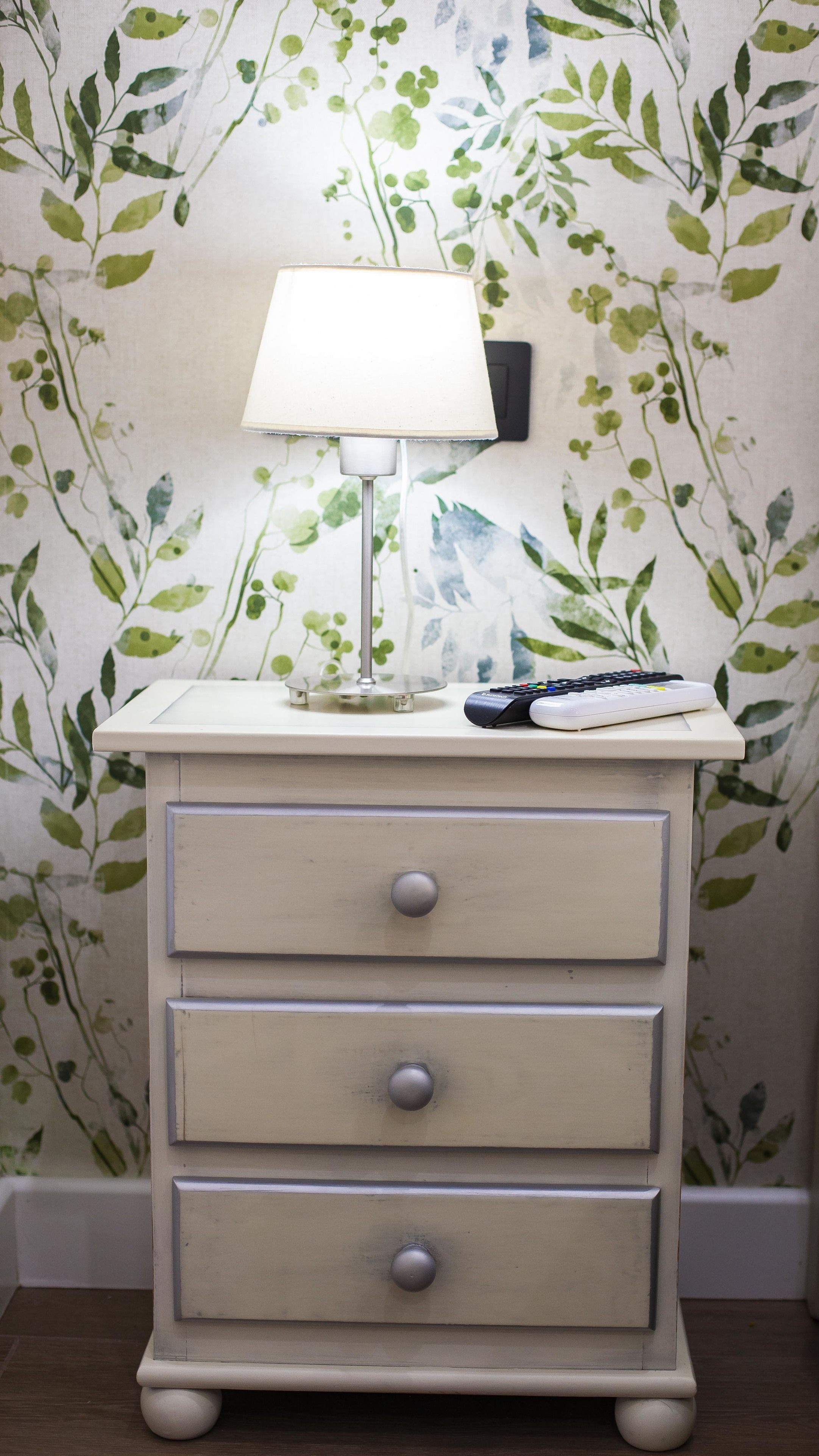 A white bedside table with a lamp and remote controls, set against a wall with a leafy wallpaper pattern.