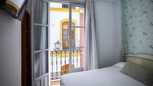 A hotel room with a bed, a window with curtains, a balcony with a view of a building, and a patterned wall. - Hostal San Francisco (Seville)