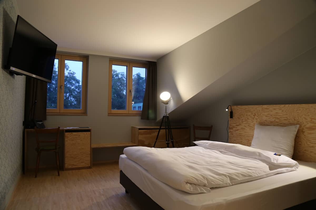 A bedroom with a bed, desk, chair, TV, and two windows.