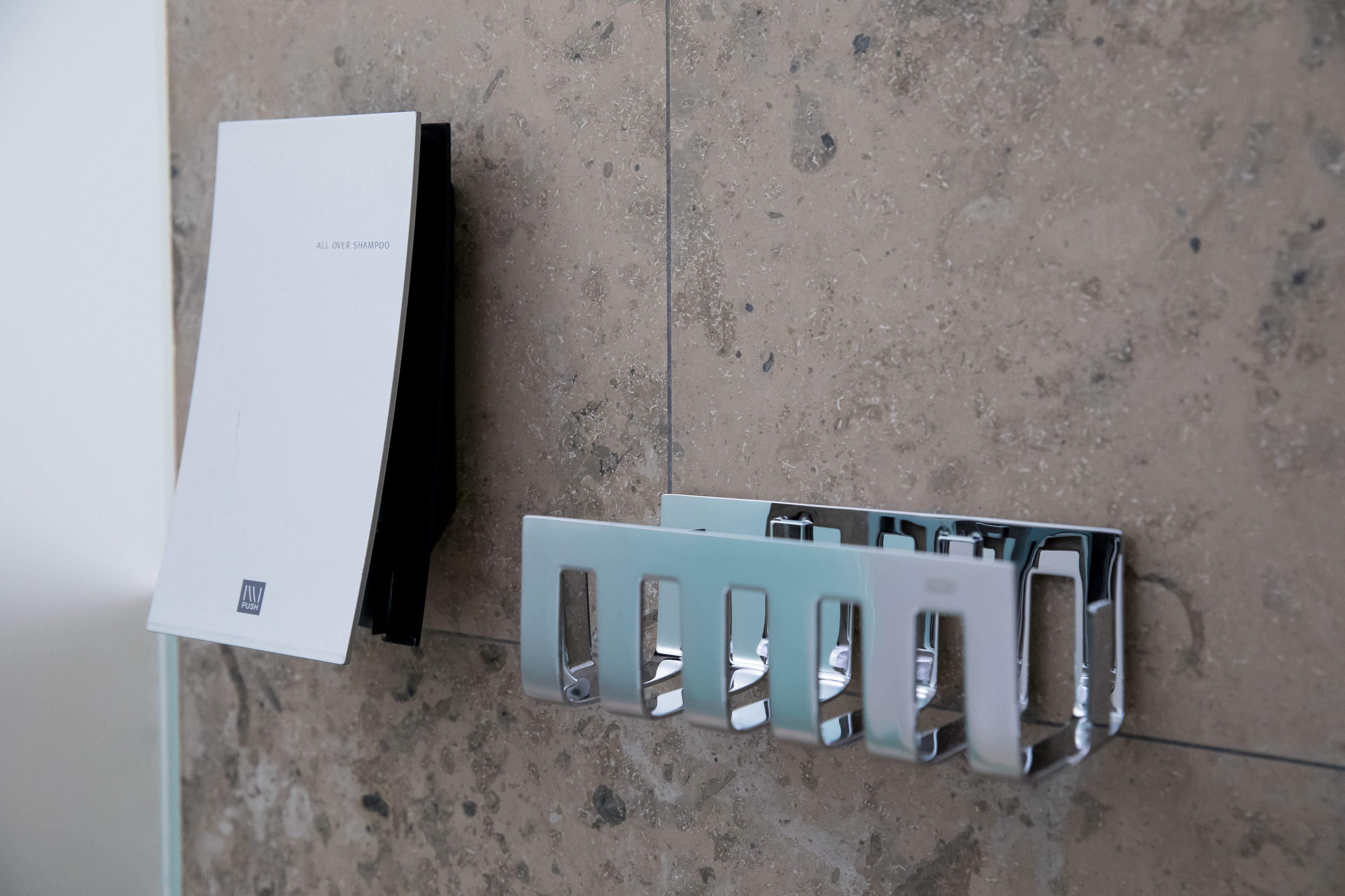 Ein Badezimmer mit weißer Wand, einem Metallhandtuchhalter und einer weißen Box mit der Aufschrift 'ALL OVER SHAMPOO'.