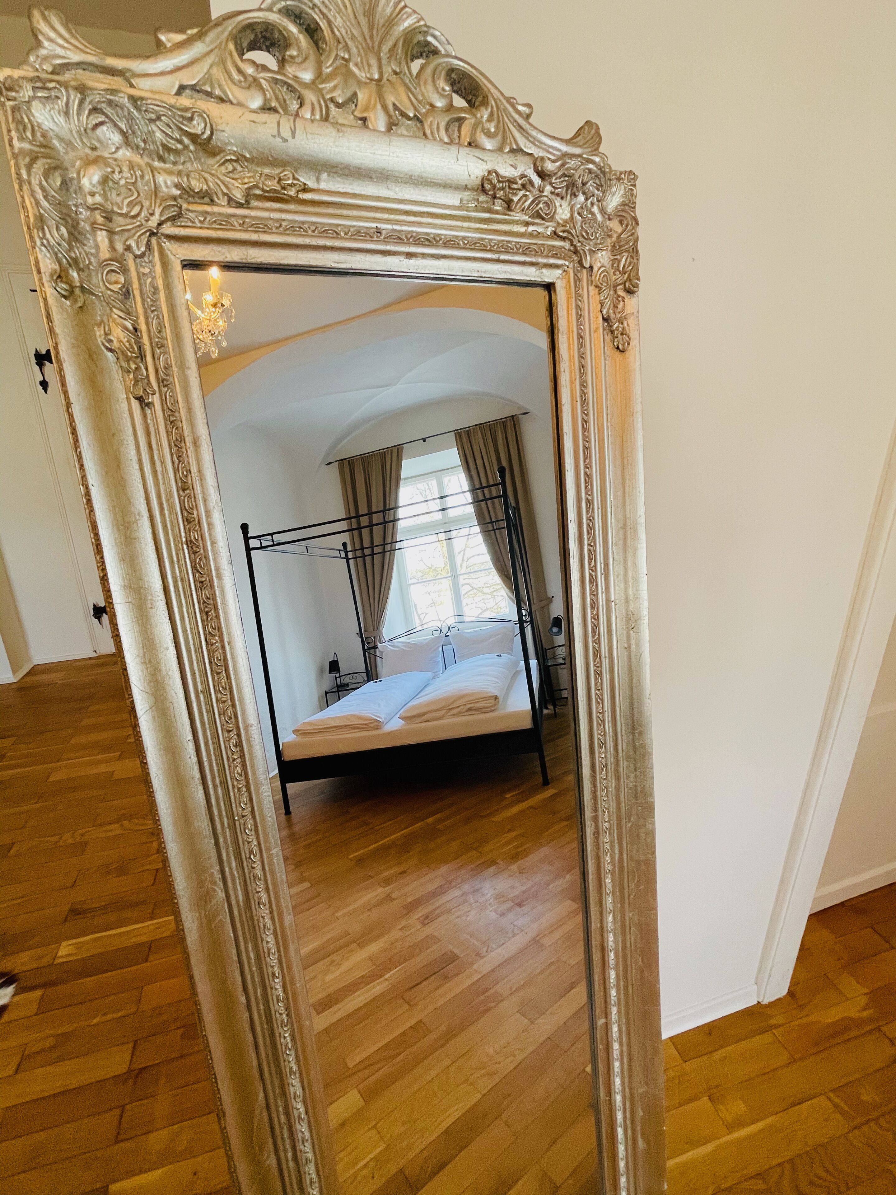 Ein Zimmer mit Holzboden, einem Bett mit Metallgestell und einem großen, verzierten Spiegel, der das Zimmer widerspiegelt.