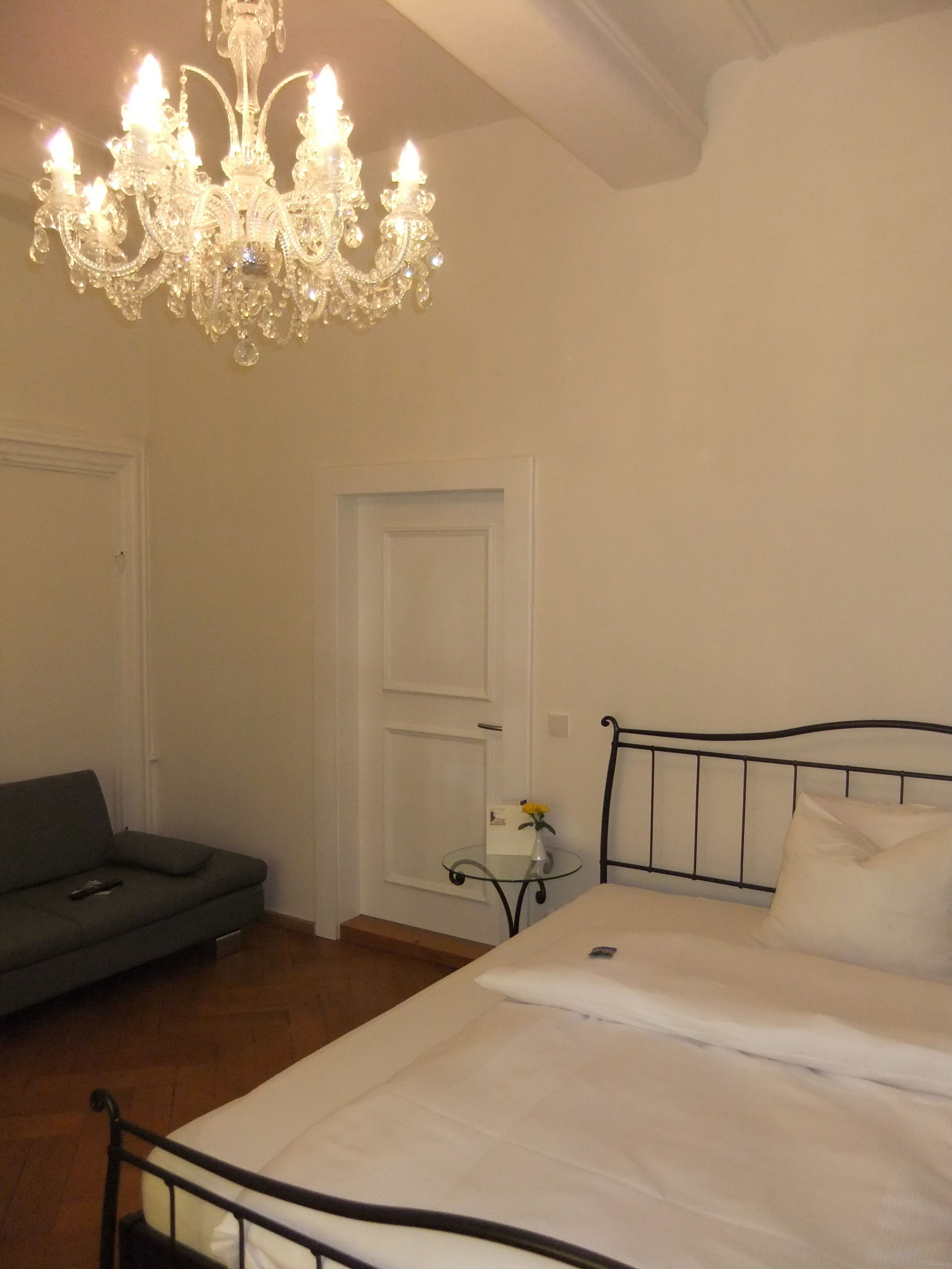 A bedroom with a bed, a grey sofa, a small table with a vase, and a chandelier.
