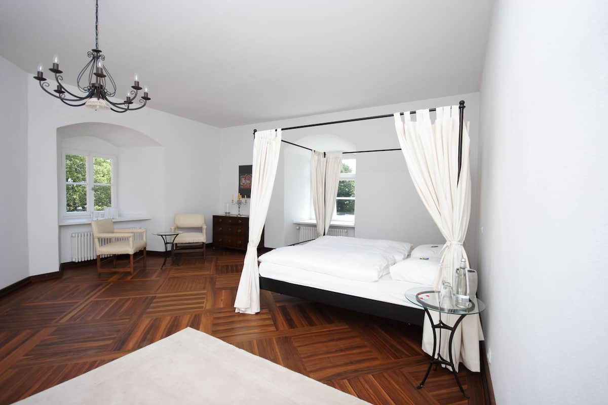 A spacious bedroom with a four-poster bed, a chandelier, and a view of the outdoors through a window.