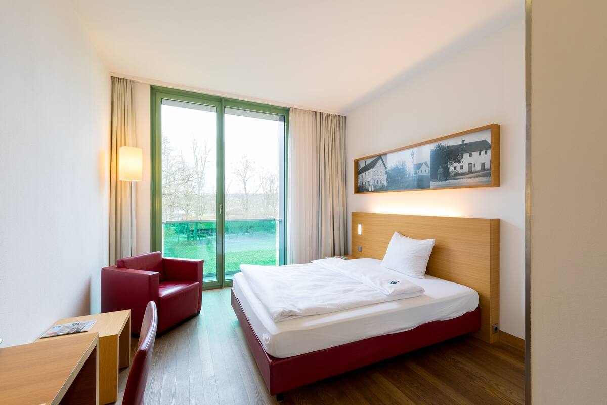 A hotel room with a bed, a desk, a red chair, a window with curtains, and a framed picture on the wall.