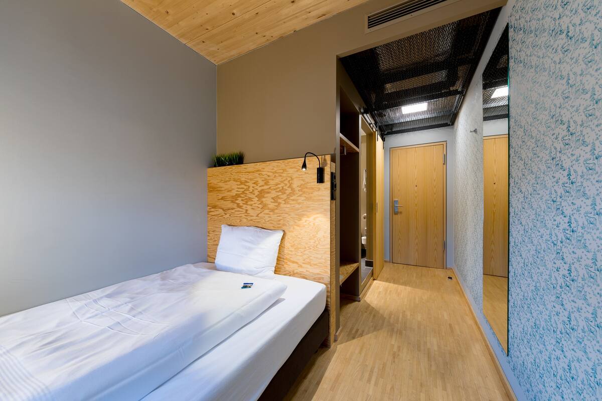 A compact room with a single bed, a wooden headboard, a small shelf, and a wardrobe.