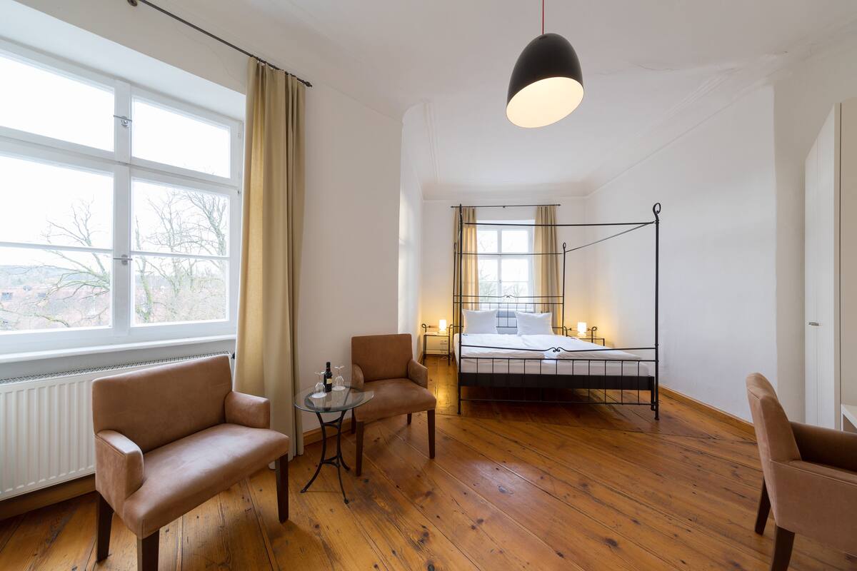 A room with a bed, two armchairs, a small table with a bottle and glasses, and a large window with curtains.