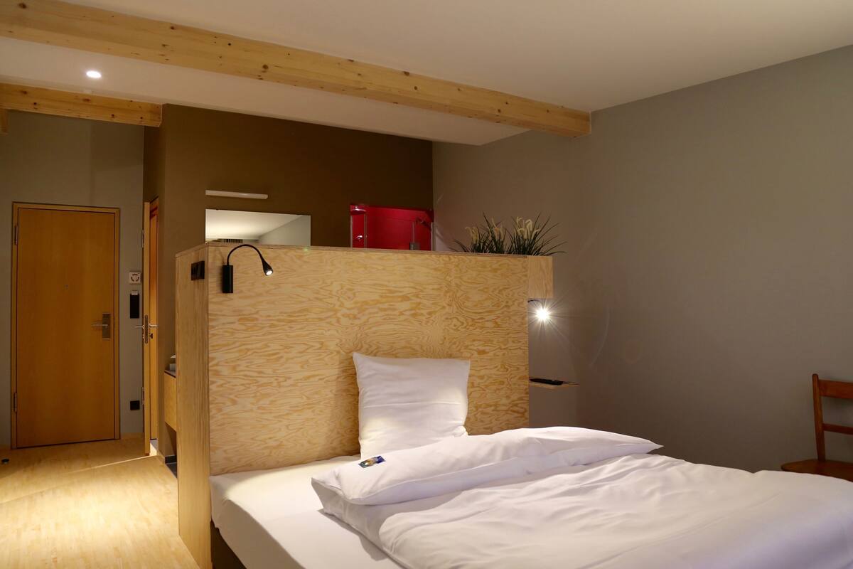 A hotel room with a wooden headboard, a bed with white linens, a red chair, and a wooden door.