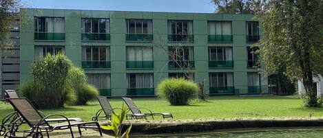 A green building with multiple windows and a small pond with lounge chairs in the foreground.