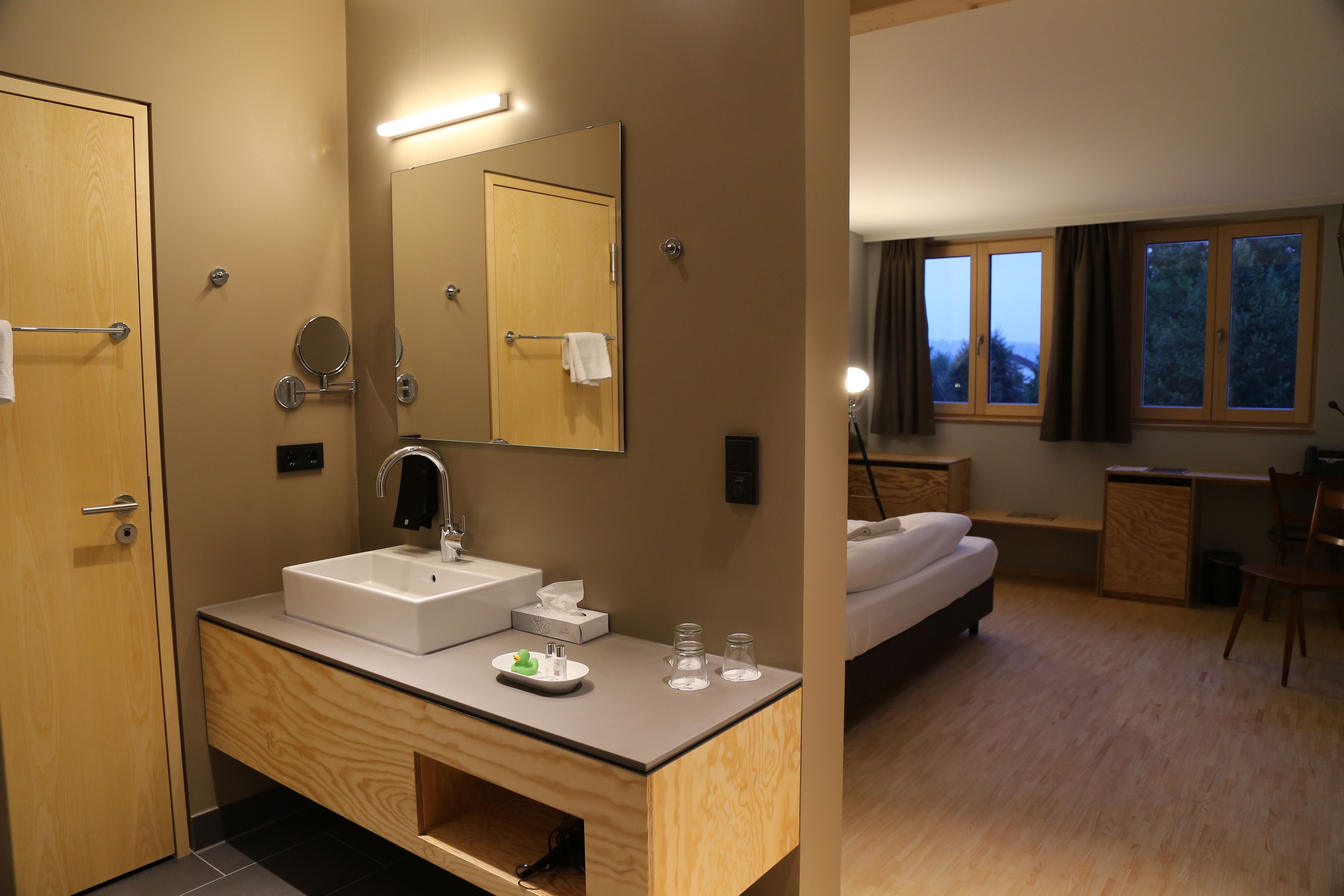 Moderni hotellihuone, jossa on puinen meikkipöytä, suuri peili, sänky, työpöytä ja ikkunasta avautuva näkymä ulos.