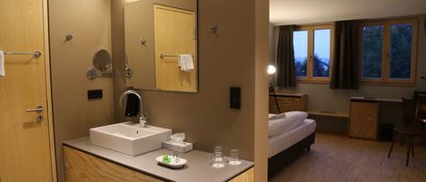 Una habitación de hotel moderna con un tocador de madera, un espejo grande, una cama, un escritorio y vistas al exterior a través de la ventana.
