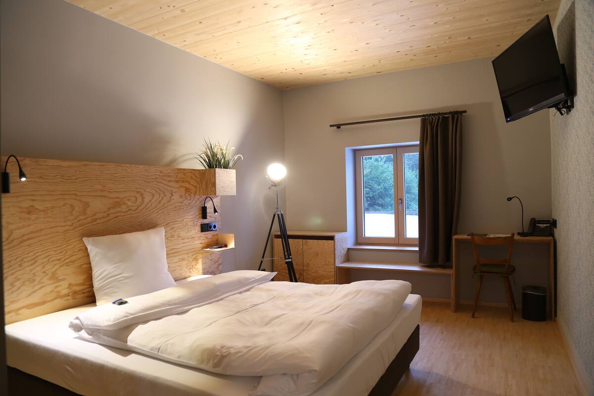 A bedroom with a wooden headboard, a bed with white linens, a desk with a chair, a TV mounted on the wall, and a window with curtains.