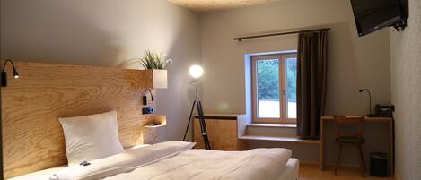 Un dormitorio con cabecera de madera, cama con ropa blanca, escritorio con silla, televisor montado en la pared y ventana con cortinas.