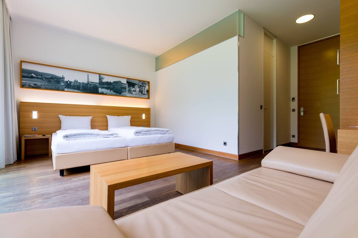 A modern hotel room with a large bed, a wooden headboard, a bedside table, a nightstand, a wooden bench, and a large wall-mounted picture.