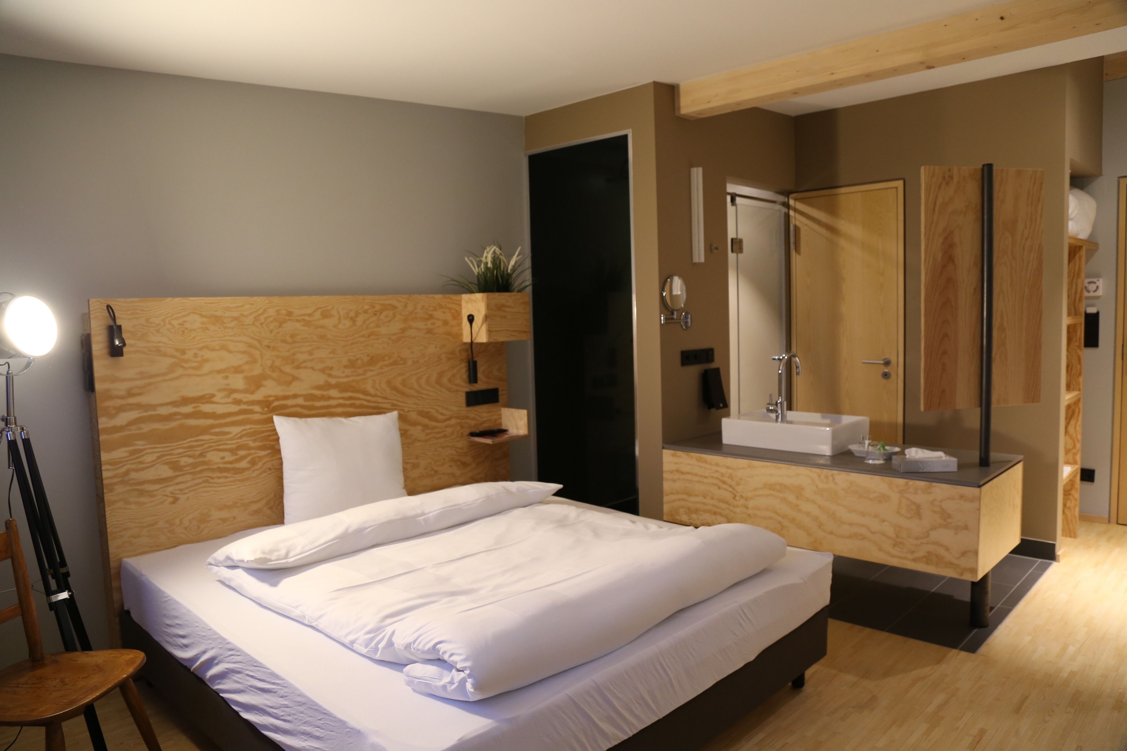 A modern hotel room with a wooden headboard, a large bed, a built-in vanity with a mirror, and a shower area.