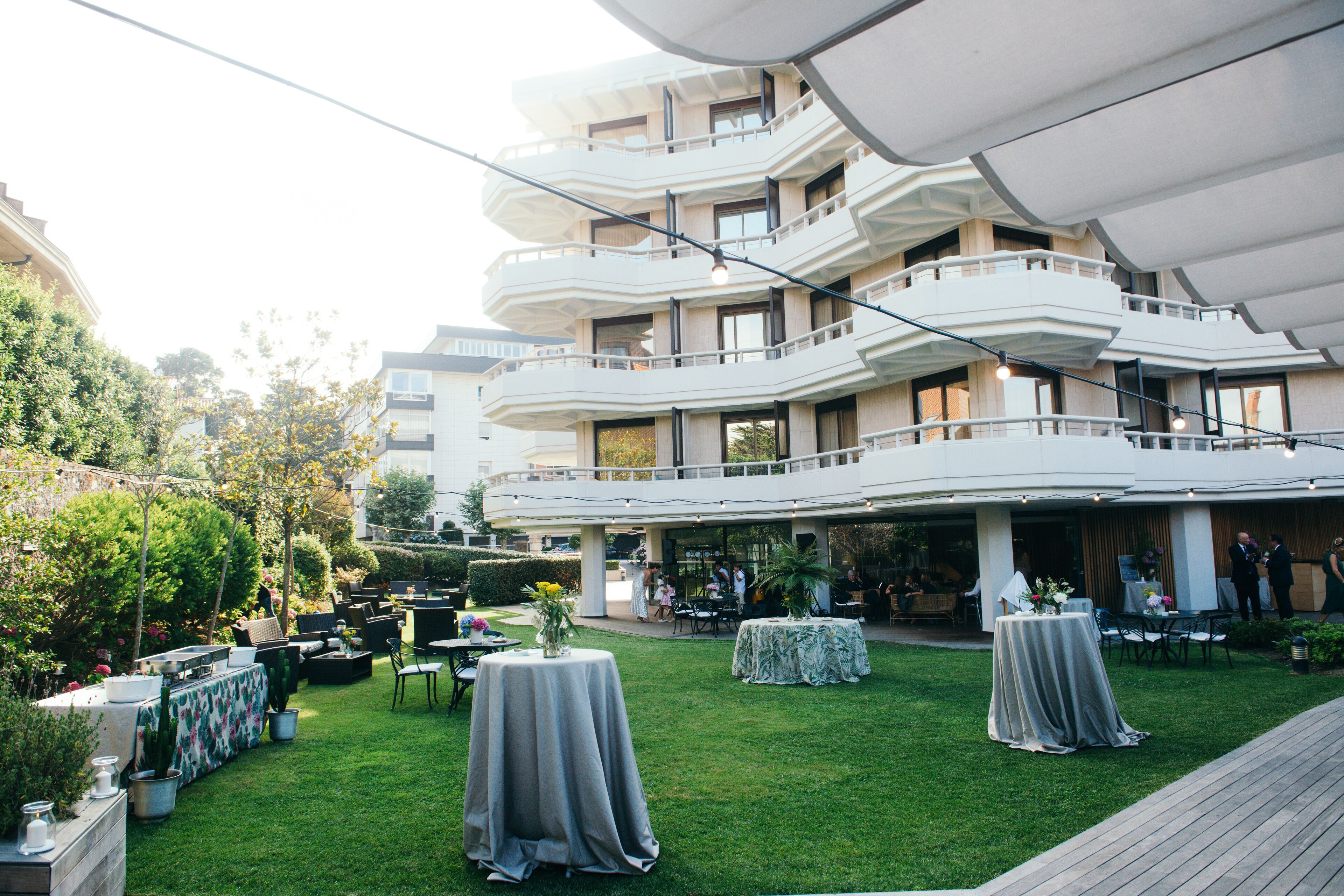 Un espacio al aire libre moderno para eventos, con varias mesas y sillas, rodeado de abundante vegetación y un edificio de varios niveles al fondo.