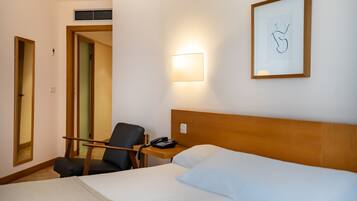 Una habitación de hotel con una cama, una silla, una mesita de noche con un teléfono, un cuadro enmarcado en la pared y una lámpara.