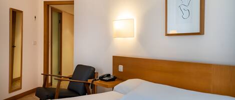 Quarto de hotel com cama, cadeira, mesa de cabeceira com telefone, quadro na parede e luminária.