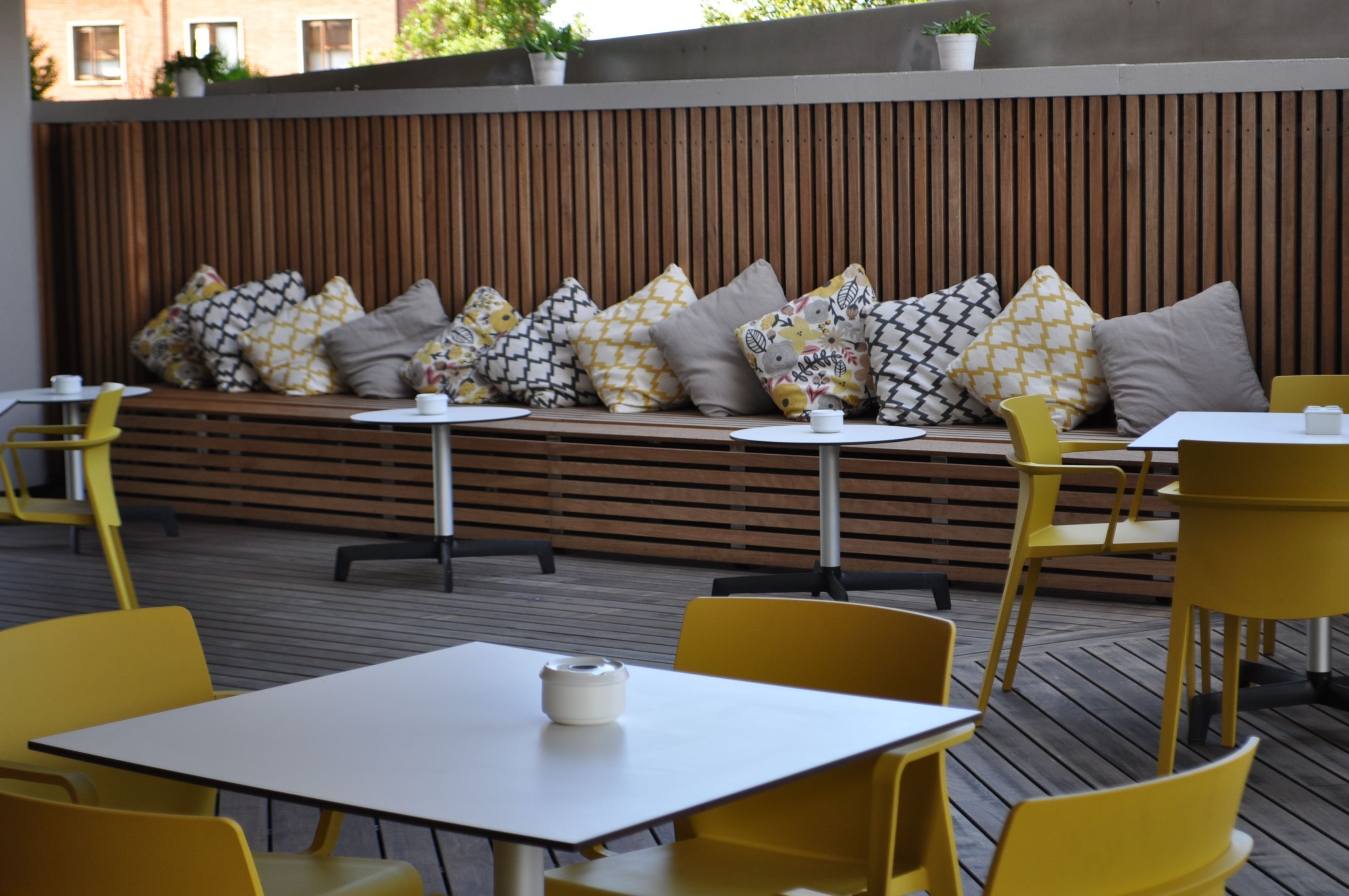 An outdoor seating area with yellow chairs and tables, a wooden slatted wall, and patterned cushions.