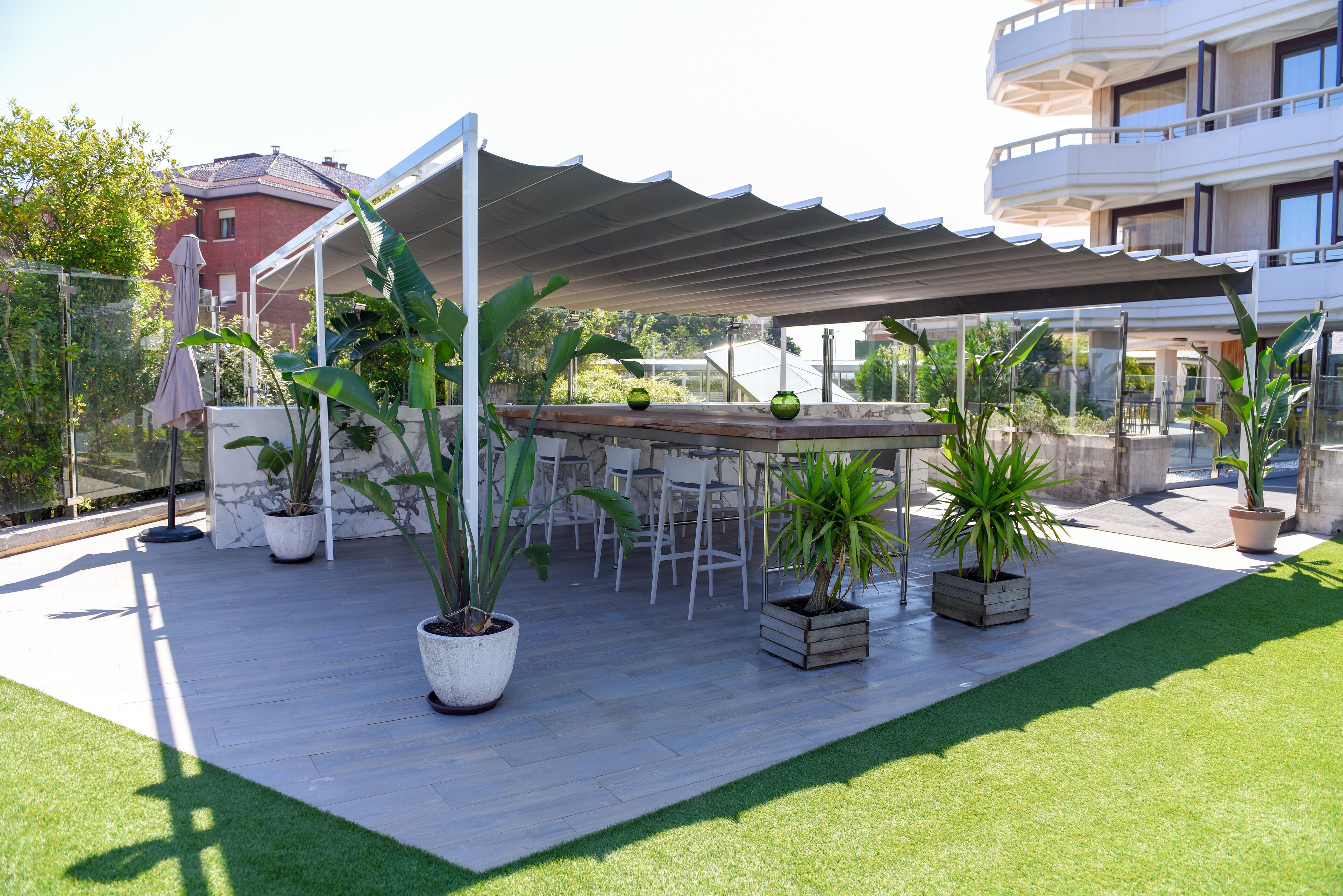 Un patio al aire libre con una zona de estar cubierta, plantas en macetas y una vista de un edificio al fondo.