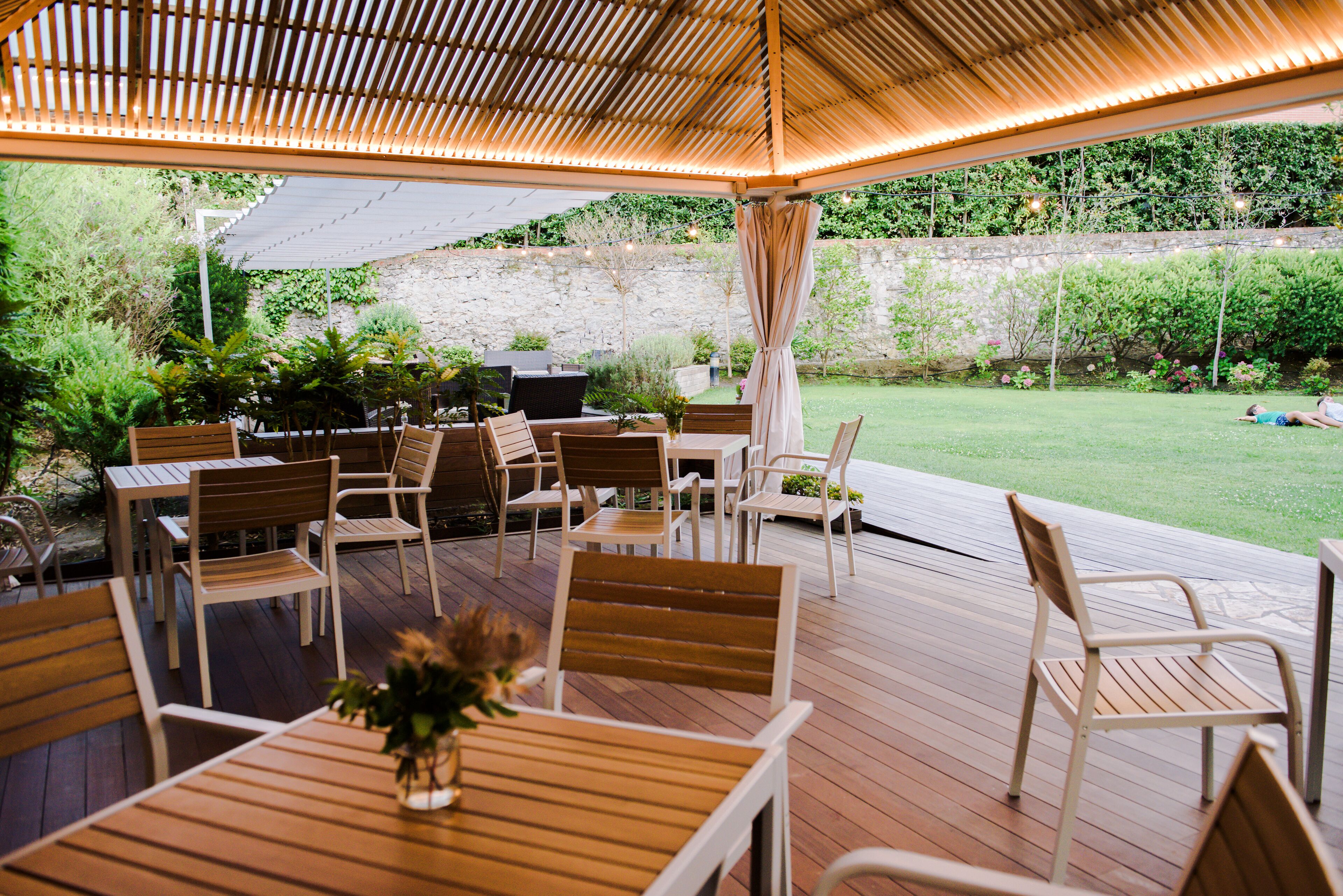 An outdoor patio with wooden tables and chairs, a grill, and a stone wall in the background.