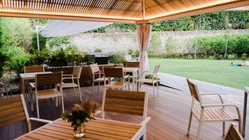 An outdoor patio with wooden tables and chairs, a grill, and a stone wall in the background.