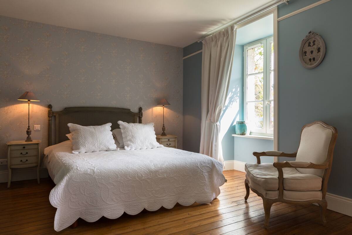 Une chambre avec un plancher en bois, un lit recouvert d’une couette blanche, deux tables de chevet avec des lampes, une chaise et une fenêtre avec des rideaux.