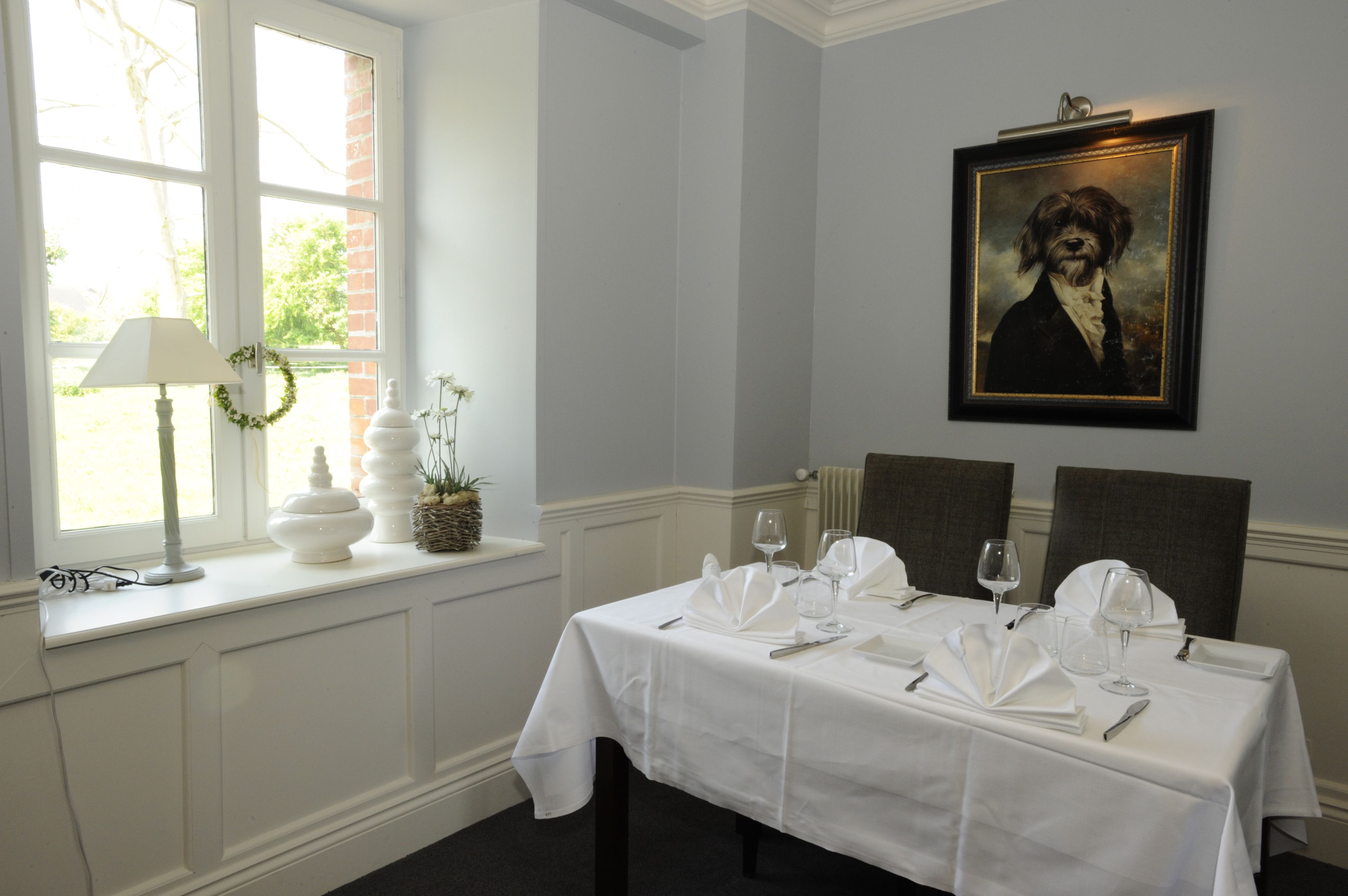 Een eettafel gedekt met een witte tafelkleed, wijnglazen en bestek, met een lijstwerkportret aan de muur.