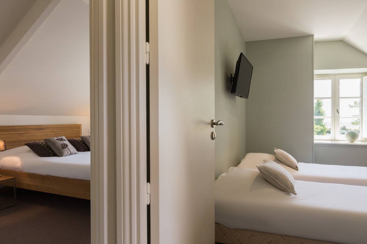 Une chambre avec un lit simple, un deuxième lit, un téléviseur fixé au mur et une fenêtre donnant sur un paysage verdoyant.