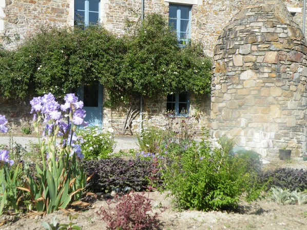 Un edificio de piedra con un jardín delantero, con flores moradas y diversas plantas.