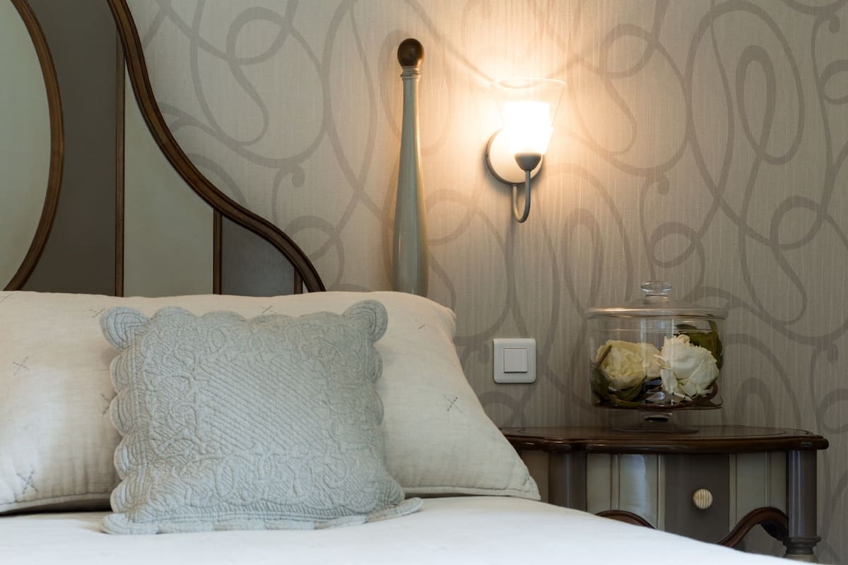 Une chambre à coucher comprenant un lit, une table de chevet, une lampe fixée au mur et un bocal décoratif posé sur la table de chevet.
