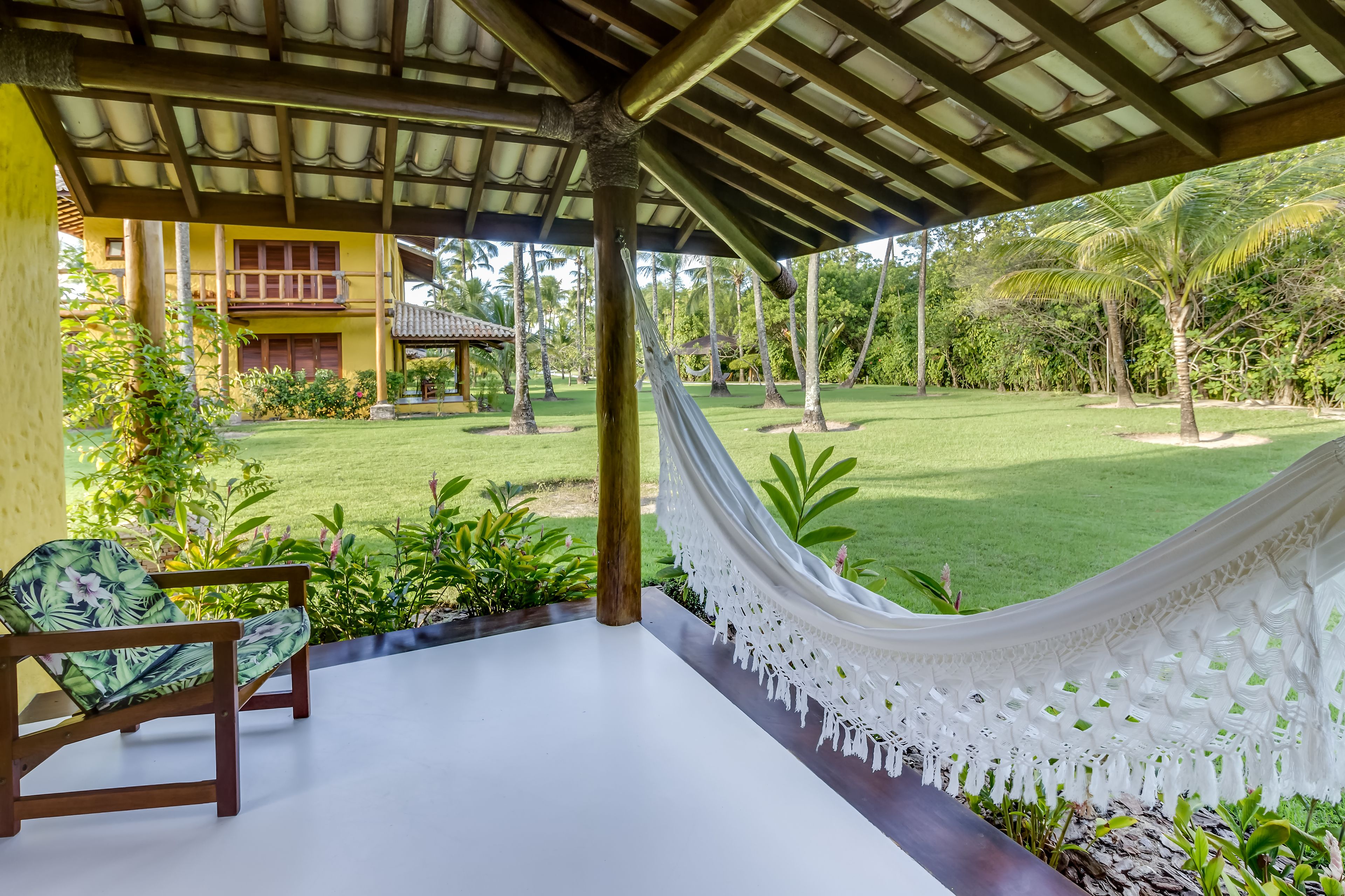 A covered outdoor area with a hammock, wooden chairs, and a view of a lush garden and a yellow building.