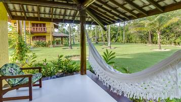 A covered outdoor area with a hammock, wooden chairs, and a view of a lush garden and a yellow building.