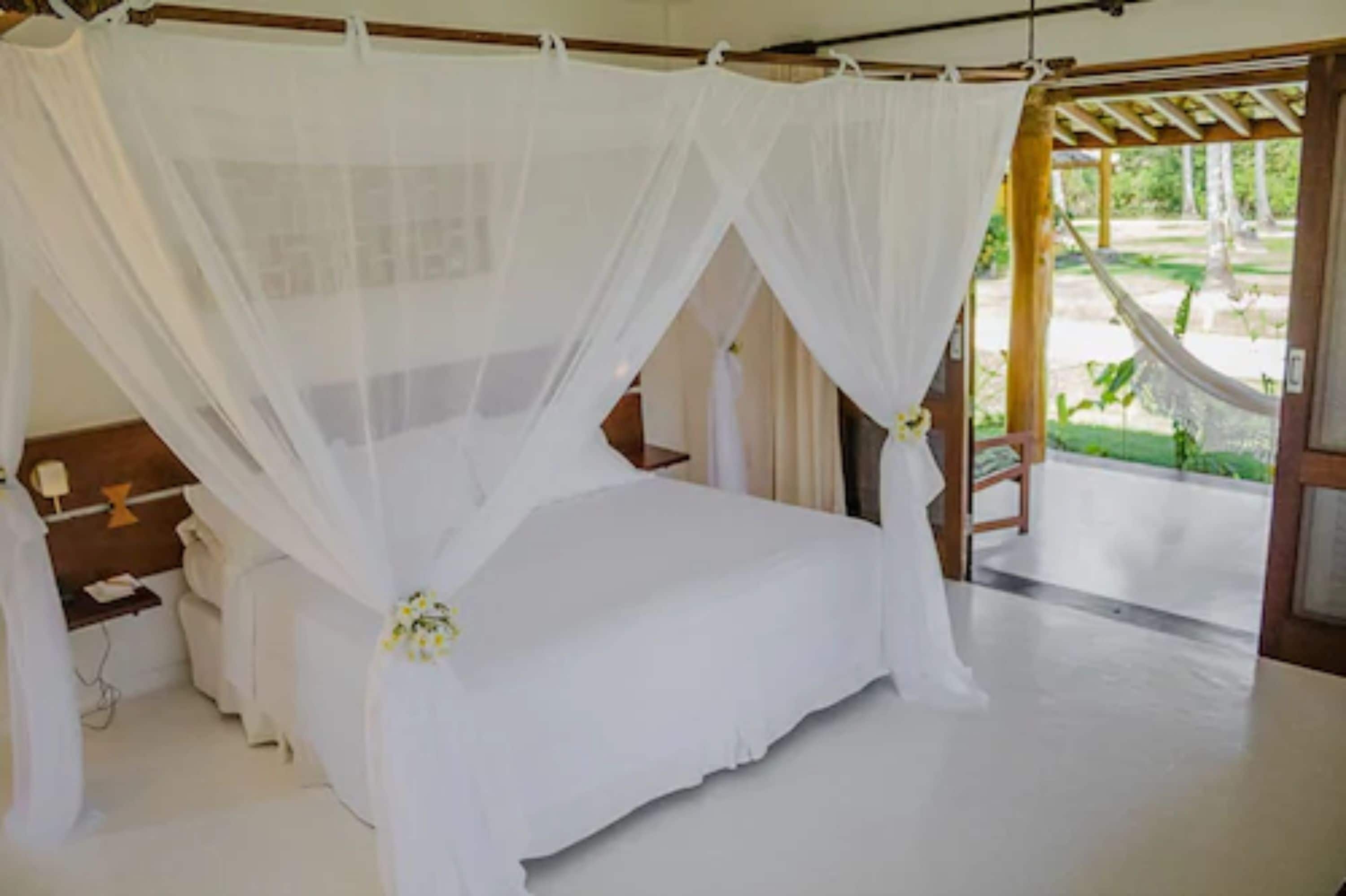 A room with a bed under a white canopy, a wooden bench, and a hammock outside.