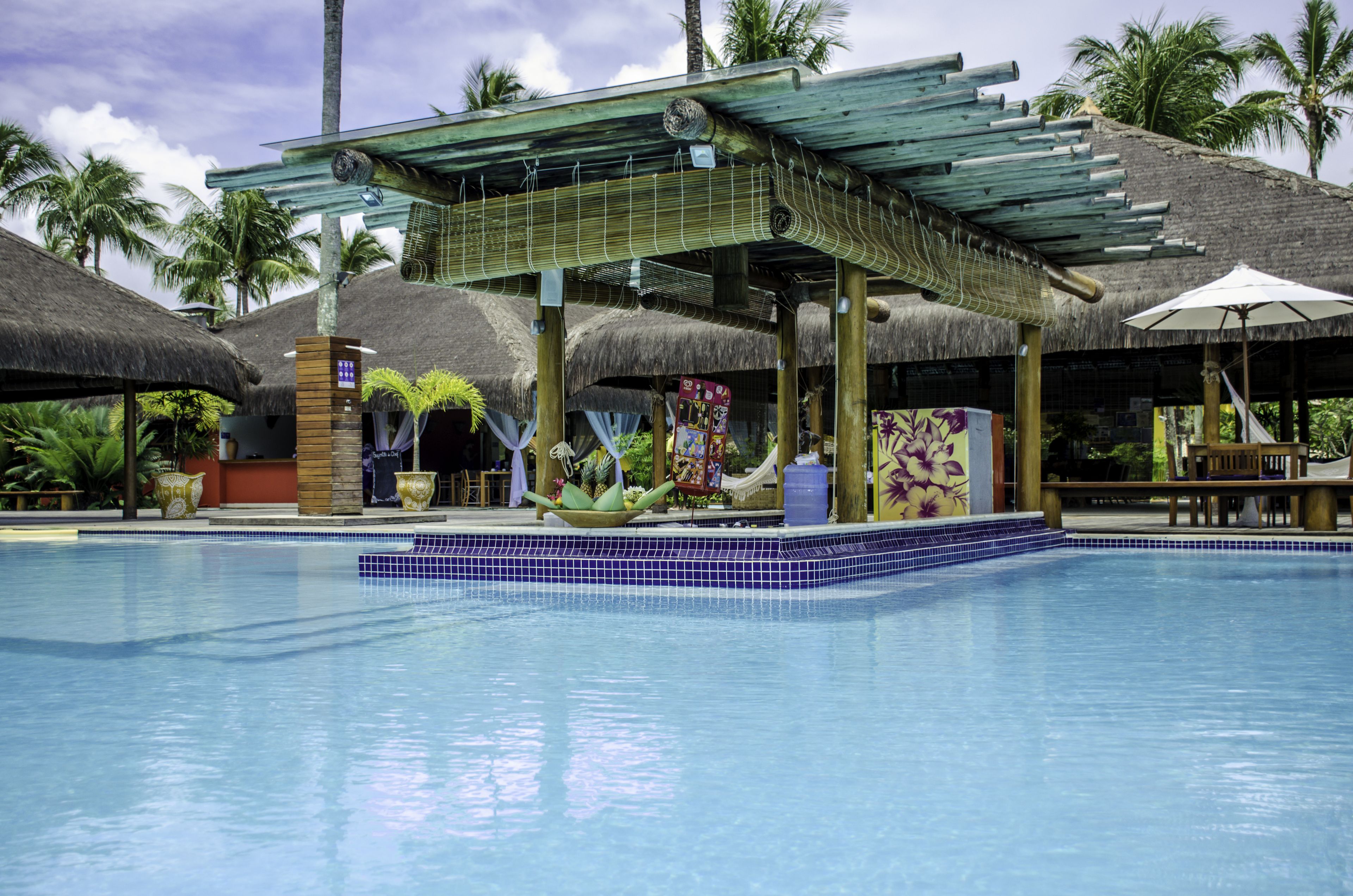 A pool area with a thatched-roof structure, palm trees, and a covered seating area.