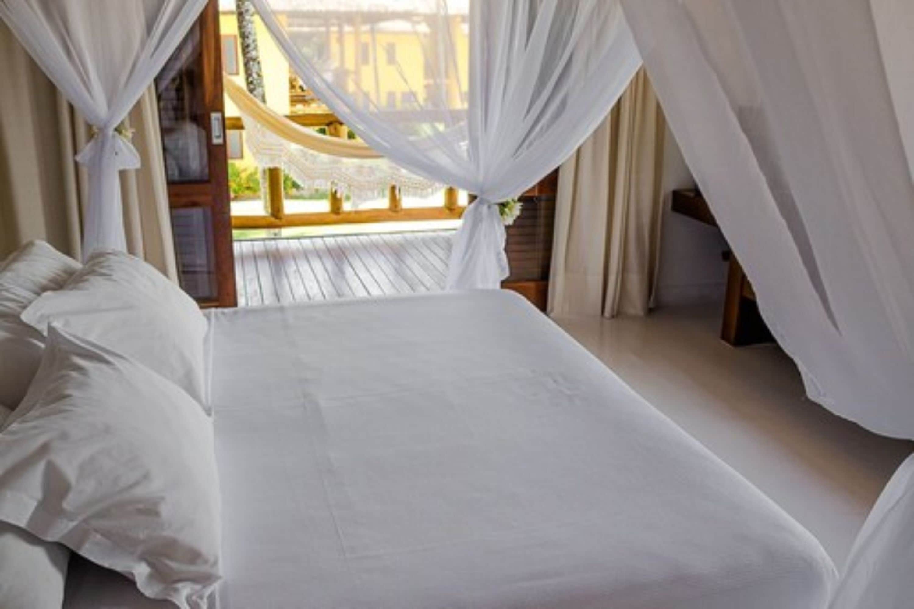 A bedroom with a large bed, white curtains, and a view of a wooden deck and building outside.