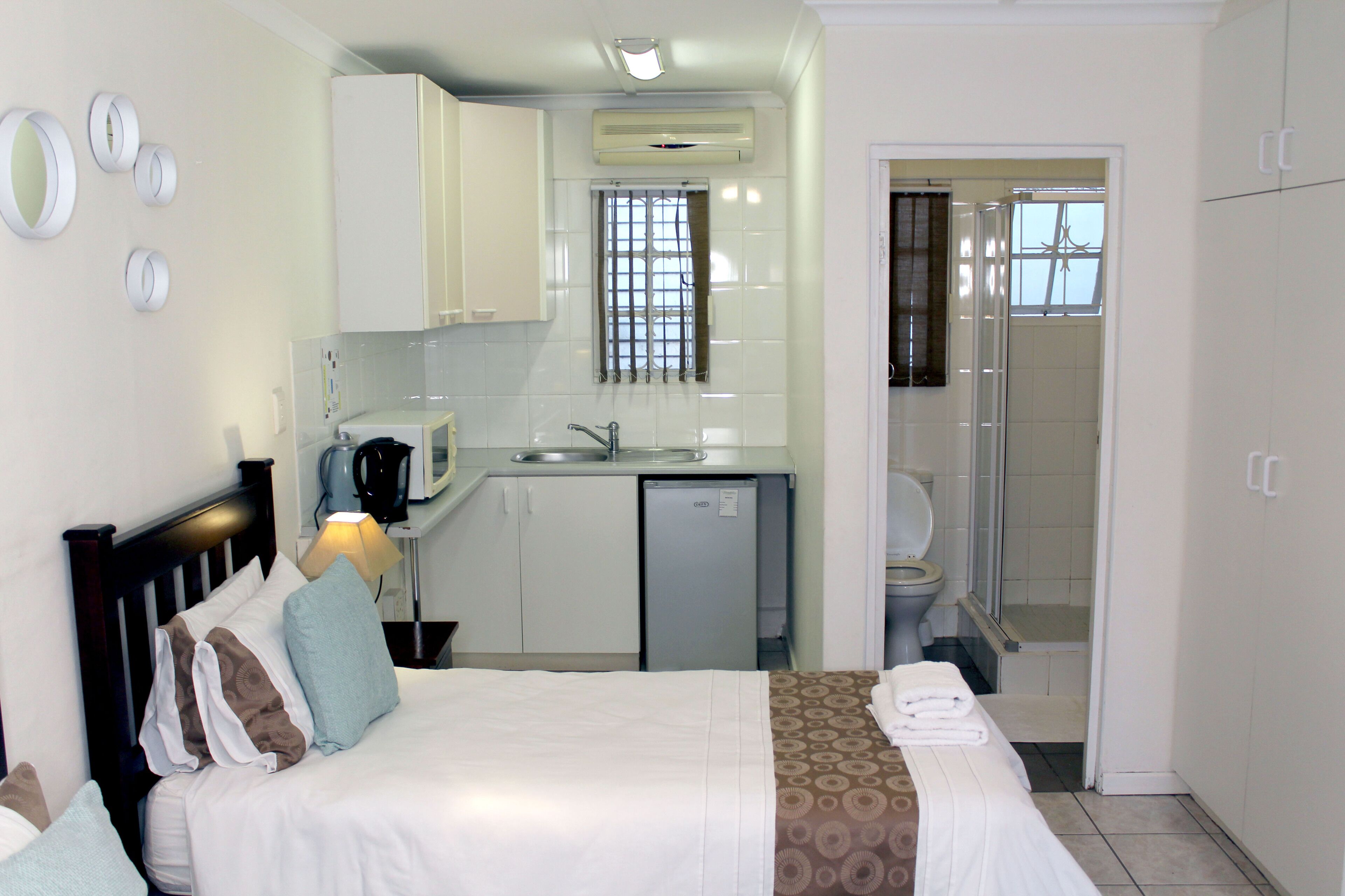 Une chambre d’hôtel de taille réduite, comprenant un lit, une petite kitchenette, une salle de bain avec douche et des toilettes.