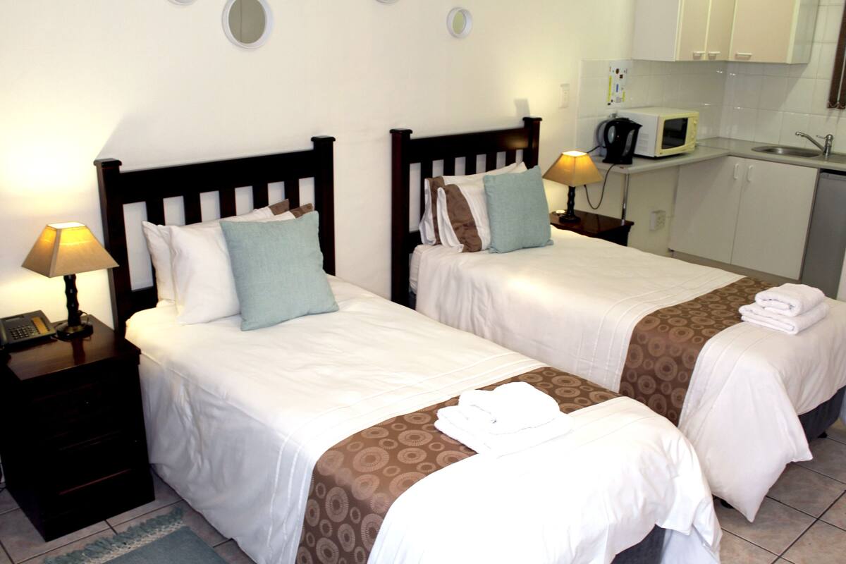 A hotel room with two beds, a nightstand with a lamp, a small desk with a telephone, and a kitchenette with a microwave and sink.