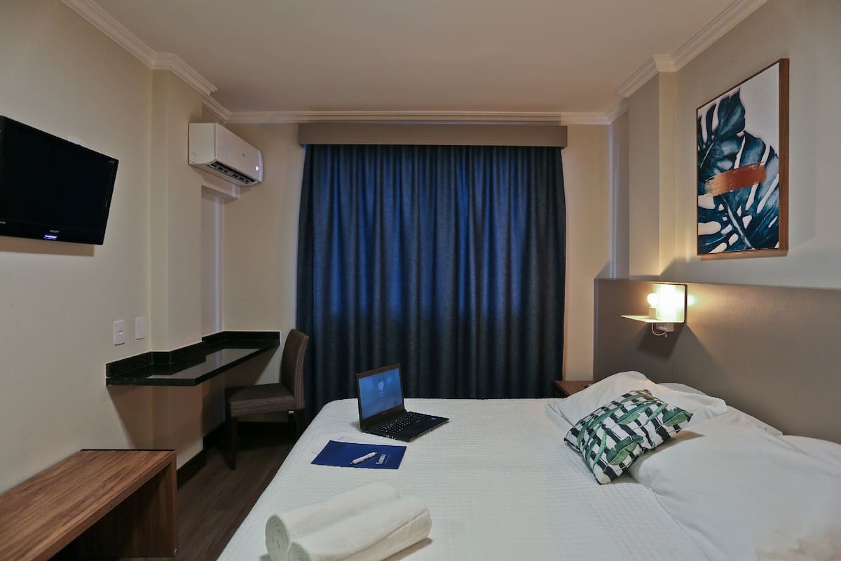 A hotel room with a bed, a desk with a laptop, a TV, and a painting on the wall.