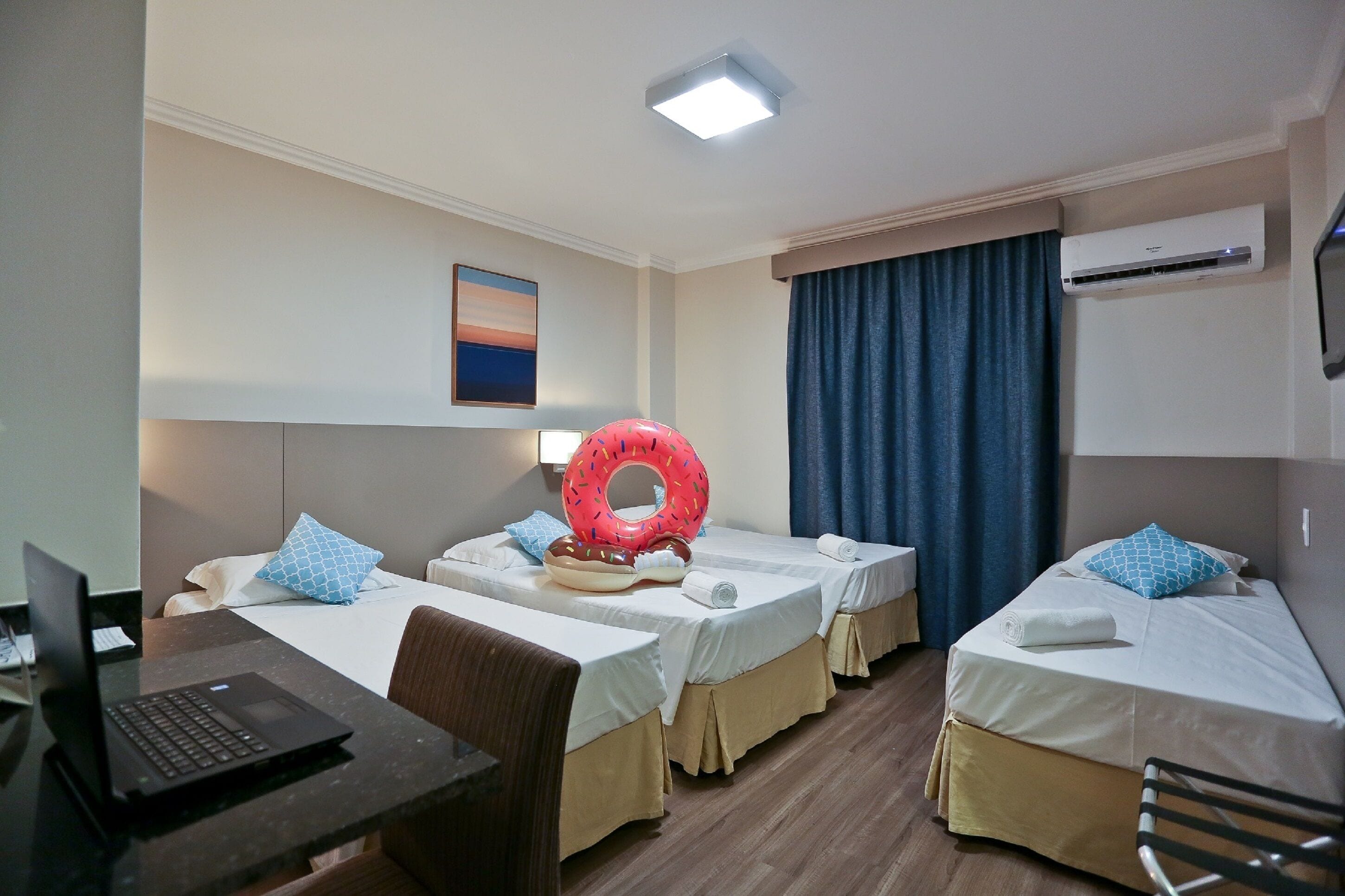 A hotel room with three single beds, a desk with a laptop, a large window with curtains, and an air conditioning unit.