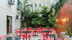 An outdoor cafe with red chairs and black tables, surrounded by lush greenery and a red wall.