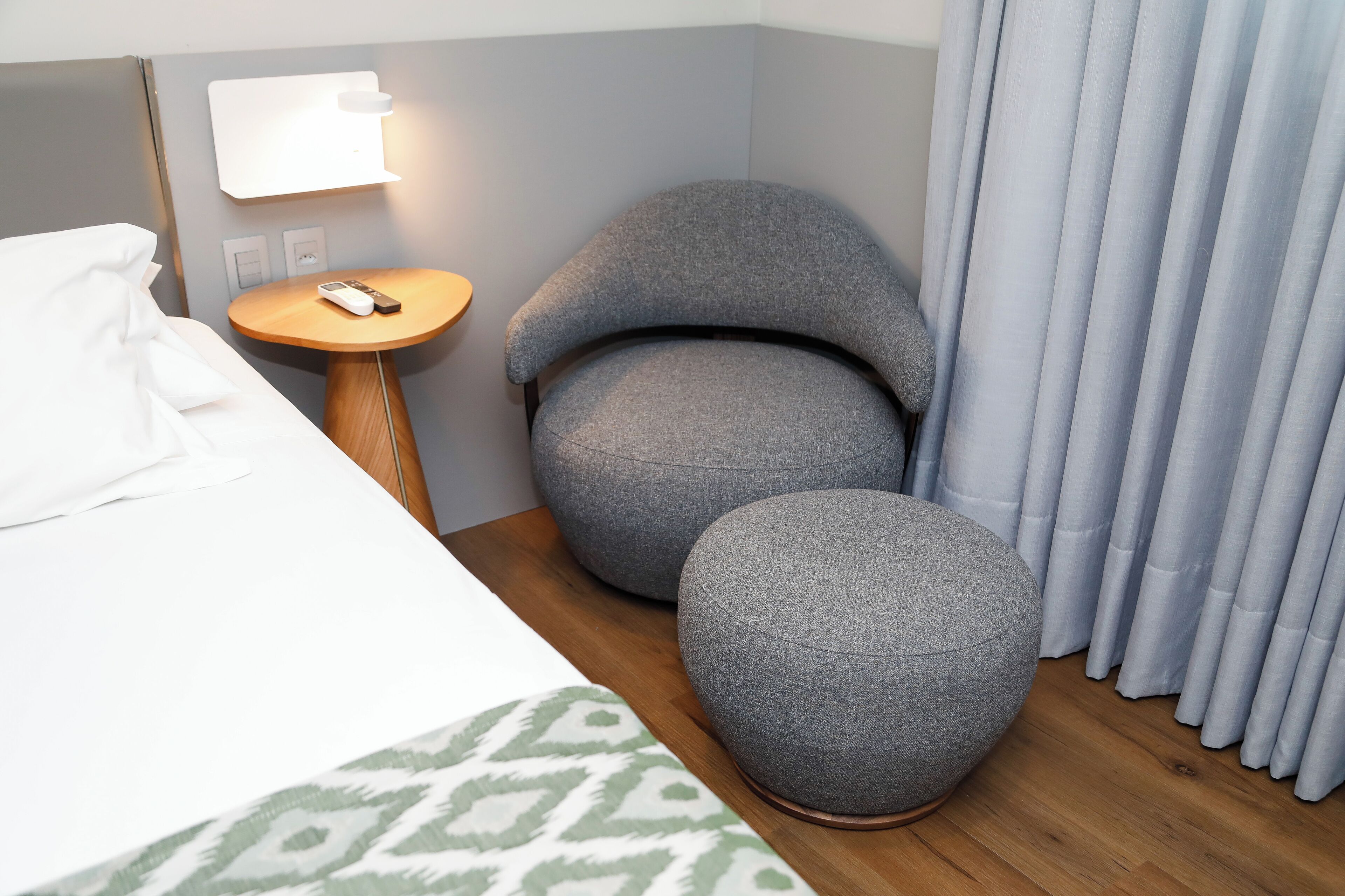 Une chambre d’hôtel avec un lit, une table ronde avec une télécommande, deux poufs gris, une lampe fixée au mur et une fenêtre avec des rideaux.