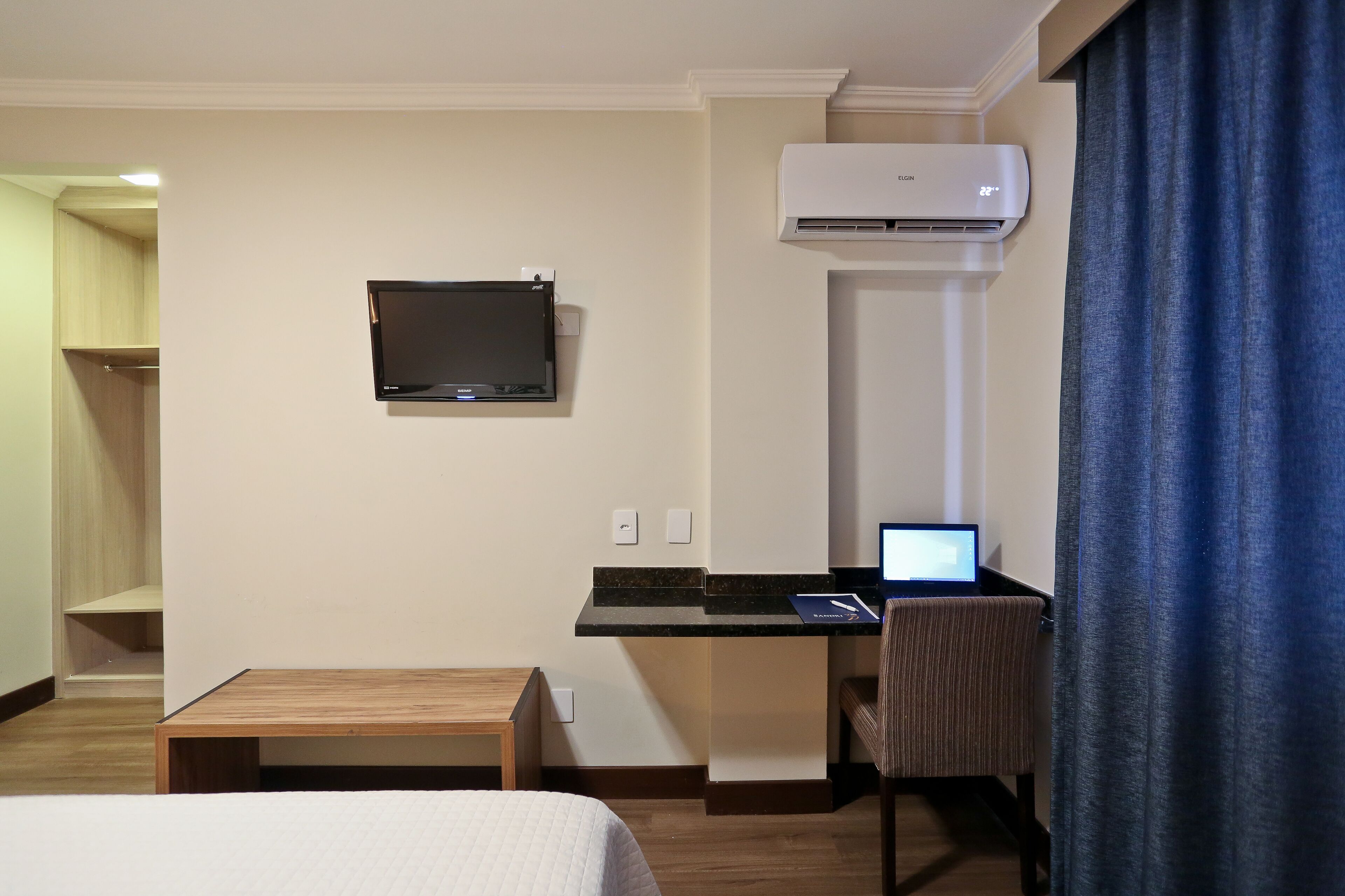 A hotel room with a bed, a desk with a laptop, a television, and an air conditioning unit.
