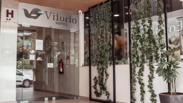 Der Eingang des Vitoria Hotel mit modernem Design, darunter eine Glastür, Zimmerpflanzen und ein sichtbarer Feuerlöscher.